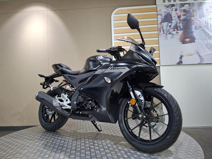 A sleek black motorcycle showcased on a circular platform, featuring a streamlined design with a prominent front fairing, sporty wheels, and modern handlebars. The backdrop includes a subtle wall with a light-colored panel and an image of people in an urban setting.