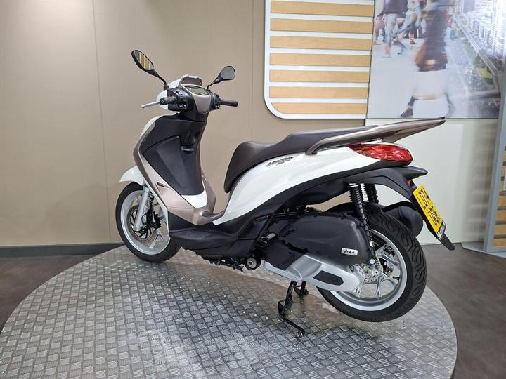 A sleek, modern scooter displayed on a circular platform with a light-colored backdrop, featuring a prominent white and gray body, clear windshield, and stylish design elements.