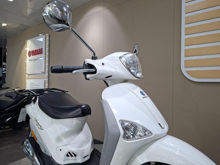 A close-up view of a white scooter with a chrome mirror, featuring a sleek design and a comfortable black seat, displayed in a showroom with a beige background.