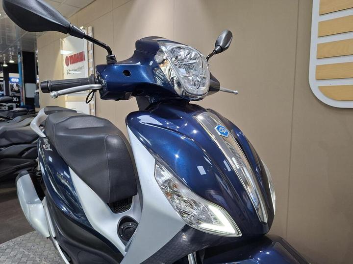 A close-up view of a blue and silver scooter, highlighting its sleek design, handlebars, headlight, and seat, set against a neutral background.