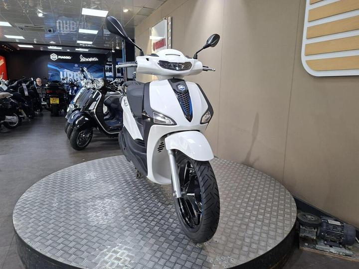 A stylish white scooter with blue accents is displayed on a circular platform in a motorcycle showroom, surrounded by various other scooters. The background features a modern, well-lit interior with signage from brands like Piaggio and Vespa.
