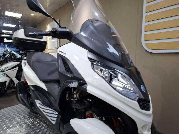 A close-up view of a white scooter showcasing its front design, windshield, and handlebar with rear-view mirrors, displayed in a showroom setting.