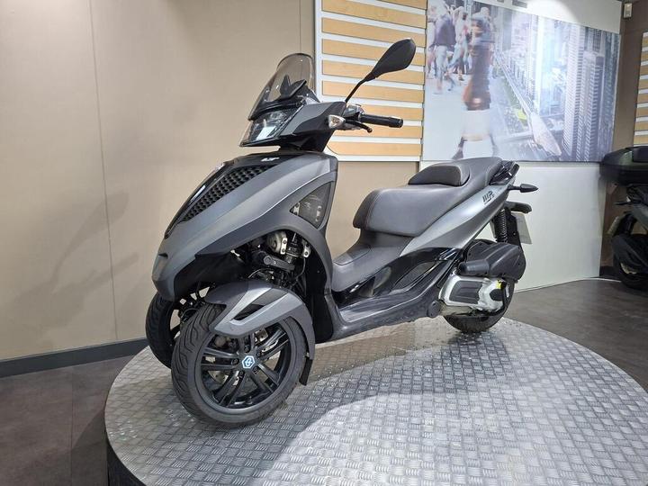 A sleek, modern three-wheeled scooter displayed on a circular platform, featuring a matte black finish, a streamlined design, and prominent front headlights. The background includes a blurred image of people walking in an urban setting.
