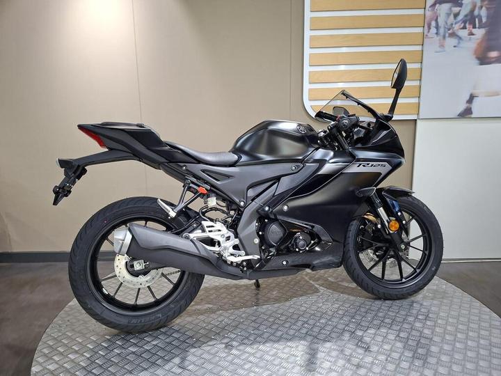 A sleek black sport motorcycle is shown from the side, featuring a modern design with sharp lines, a low profile, and sporty details. The bike is positioned on a textured display surface against a neutral background.
