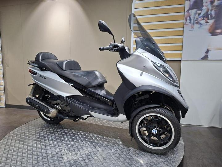 A modern silver and black scooter with a large windscreen and two seats, displayed on a circular metallic platform against a neutral wall background.