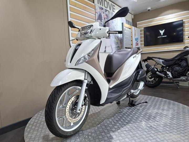 A white and brown scooter is displayed on a textured platform in a showroom, with a television screen and another motorcycle visible in the background.
