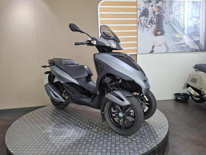 A sleek, gray three-wheeled scooter displayed on a circular platform, set against a neutral background with a promotional image of a city scene on the wall.