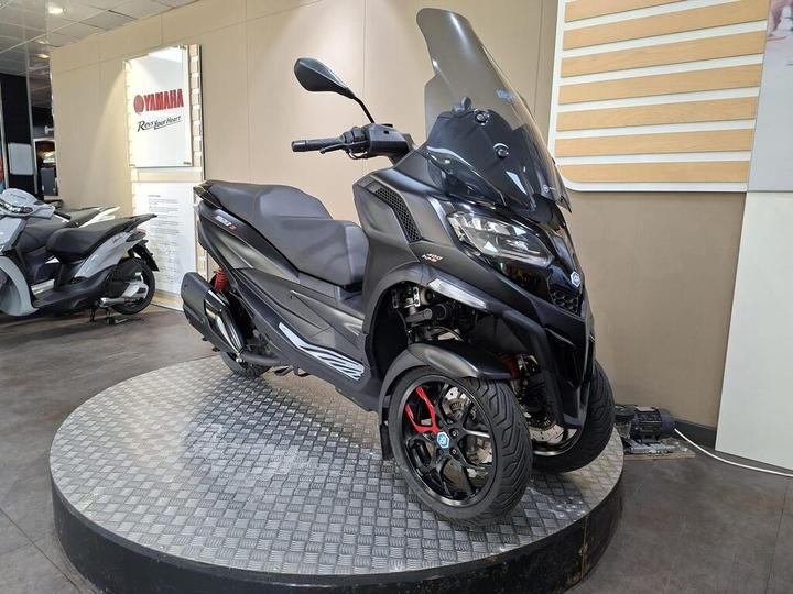 A black three-wheeled scooter displayed on a circular platform in a Yamaha showroom, featuring a large windshield and sporty design elements. A second scooter is partially visible in the background.