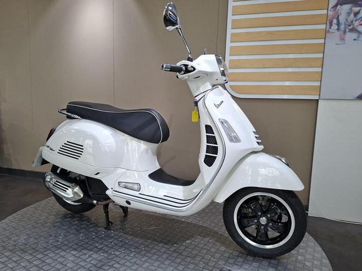 A white Vespa scooter displayed in a showroom, featuring a classic design with a black leather seat, chrome accents, and sleek wheels. The background includes a simple, modern interior with wooden paneling.