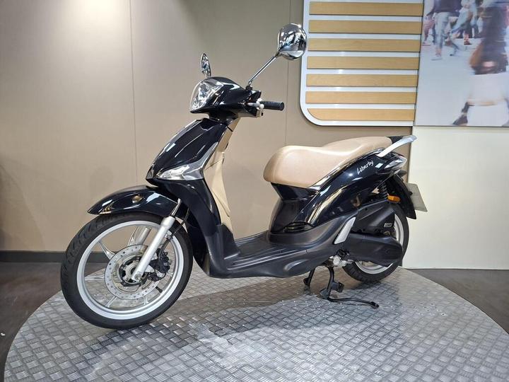 A sleek black scooter with a tan seat is displayed on a circular, textured platform against a neutral background. The scooter features a modern design with chrome mirrors and is angled to show its profile.