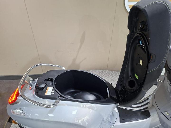 An open storage compartment on a gray scooter, with visible interior space and a circular cap, set against a neutral background.