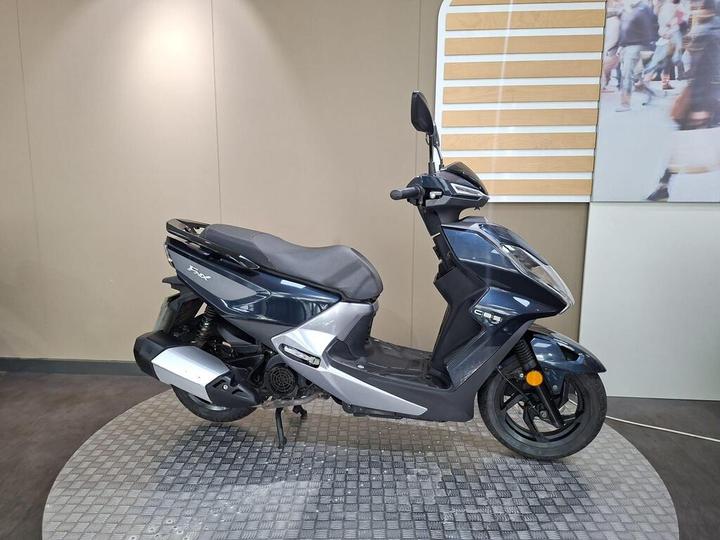 A dark blue and silver scooter is positioned on a textured circular platform against a neutral wall background.