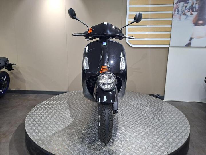 A black scooter displayed on a circular metallic platform, facing forward with a clear view of its front light, handlebars, and dashboard. The background features a neutral wall with a wooden accent panel.
