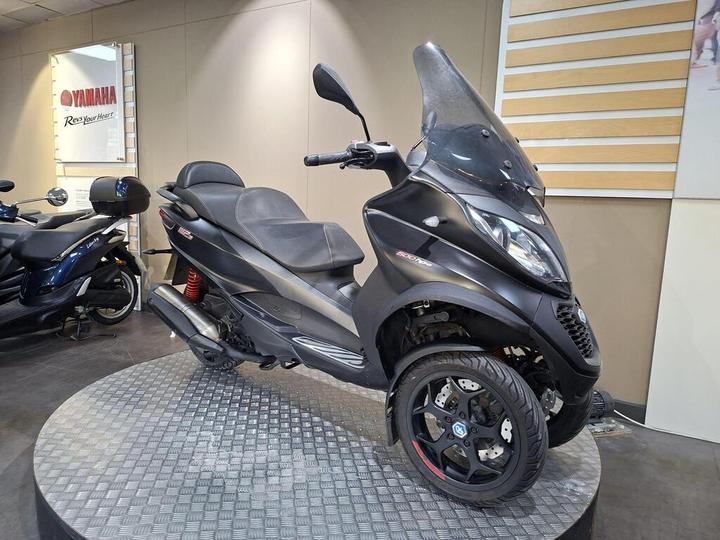 A black three-wheeled scooter displayed on a circular showcase platform in a Yamaha dealership, featuring a large windshield, sporty design, and a visible logo on the front.