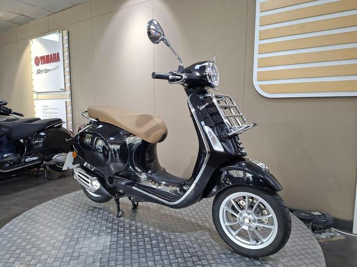 A black Vespa scooter with a brown seat is displayed on a checkered platform in a showroom, with a Yamaha sign visible in the background.