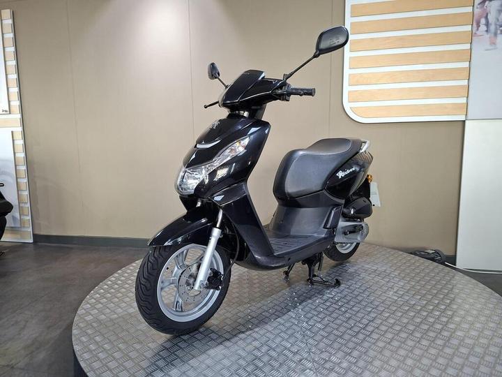 A black scooter is displayed on a circular platform with a textured surface. The scooter features a sleek design, front headlights, and a comfortable seat, set against a neutral background.