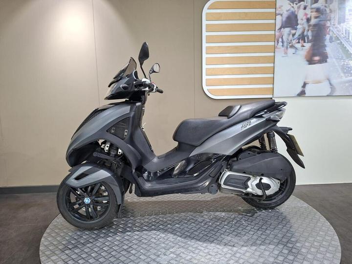 A sleek black scooter displayed on a circular textured platform, set against a neutral background with a partial image of a busy street in the upper corner.