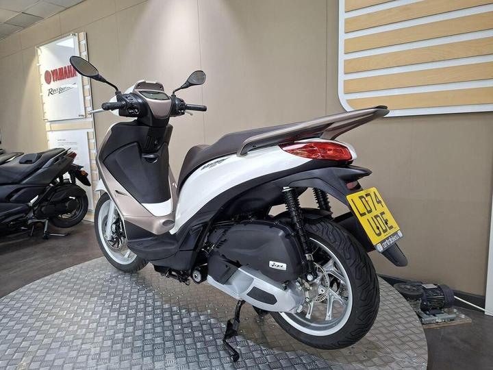 A silver and black scooter is displayed in a showroom with a modern design. The scooter is positioned at an angle, showcasing its rear and side. In the background, there is a sign for Yamaha and another motorcycle. The showroom features a textured floor.