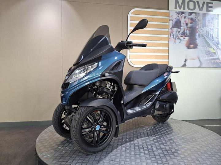 A three-wheeled blue scooter displayed on a rotating platform in an indoor setting with a modern backdrop featuring a graphic that says "MOVE."