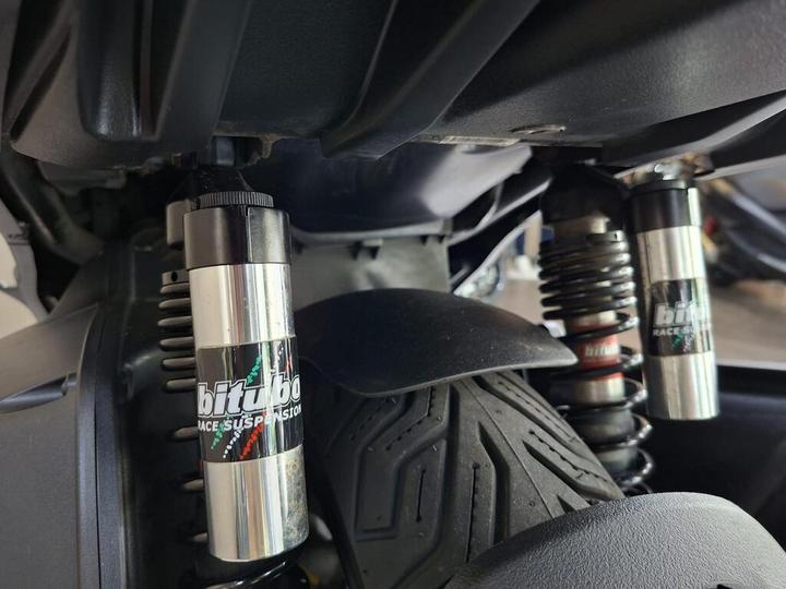 Close-up view of race suspension components, including two silver shock absorbers with branding, mounted beneath a vehicle. The background features part of a tire and the vehicle's undercarriage.