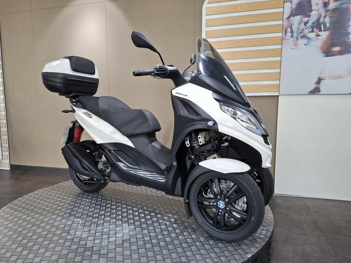 A stylish three-wheeled scooter in white and black, positioned on a circular display platform, featuring a rear storage compartment and modern design elements. The background consists of a beige wall with a decorative panel.