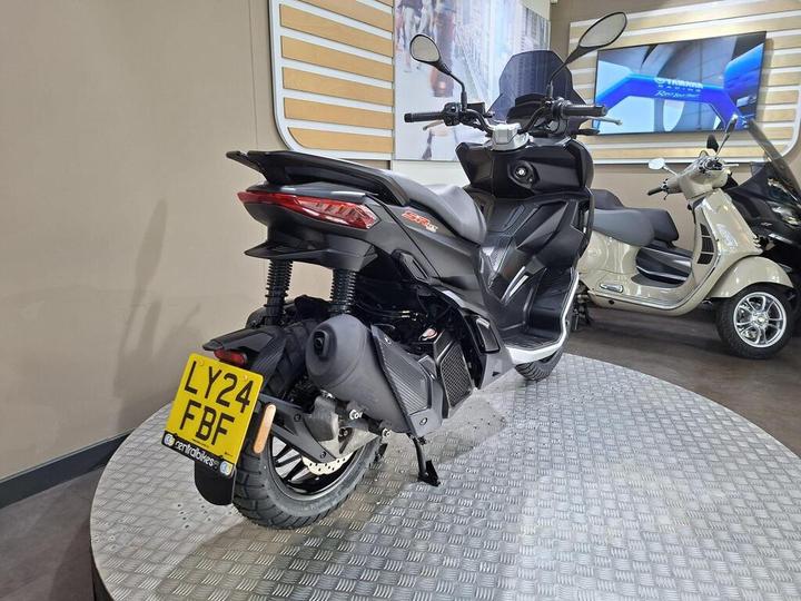 A black scooter is showcased on a circular metal platform with a checkered pattern, featuring a visible license plate marked "LY24 FBF." Behind it, another scooter in a cream color can be seen, along with a wall displaying digital screens.