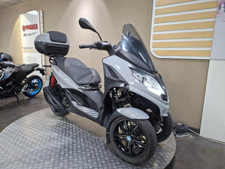 A gray three-wheeled scooter with a high windshield and a storage compartment on the back, displayed on a textured circular platform in a showroom, with other motorcycles visible in the background.