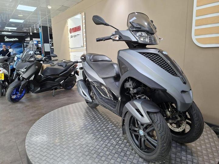 A display of two scooters in a showroom; the foreground features a gray three-wheeled scooter with a sleek design, while a dark two-wheeled scooter is partially visible behind it. The showroom has a modern aesthetic with bright lighting and promotional signage.