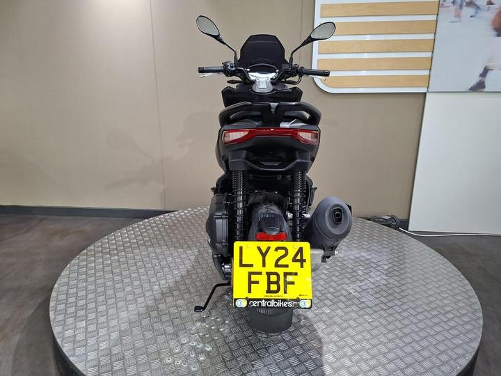 A rear view of a motorcycle on a textured circular platform, showcasing the tail section, license plate with the number "LY24 FBF," and side exhaust. The background is minimal with a light-colored wall.