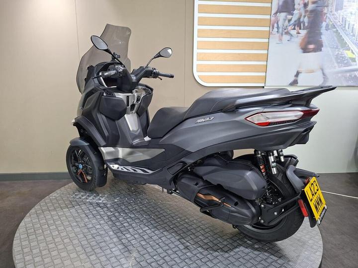 A sleek black three-wheeled scooter displayed on a textured platform, with a clear windshield and modern design elements, against a minimalist backdrop.