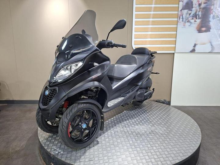 A black three-wheeled scooter displayed on a circular platform, featuring a windshield and sporty design elements. The background shows a neutral-colored wall with a partially visible image of people walking.
