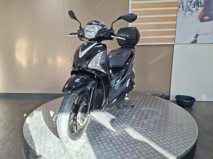 A black scooter displayed on a circular platform with a light background and sunlight streaming in.
