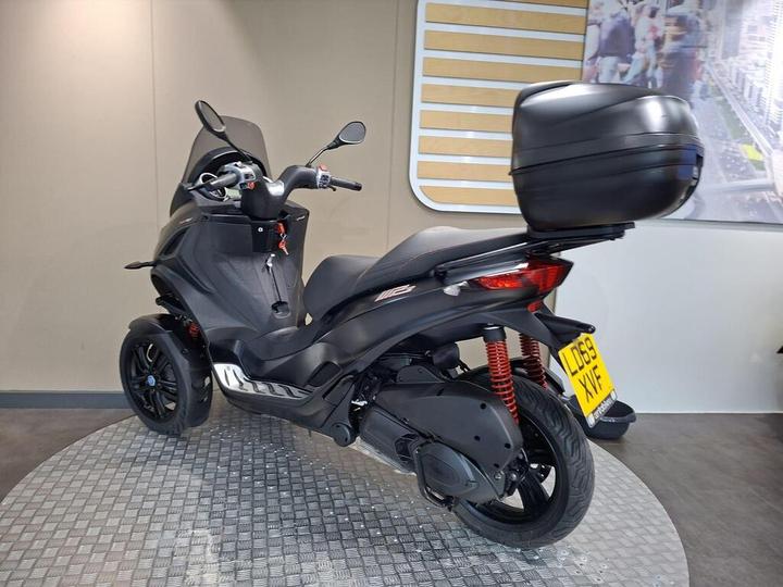 A sleek black three-wheeled scooter with a rear storage case is positioned on a circular platform against a minimalist backdrop.
