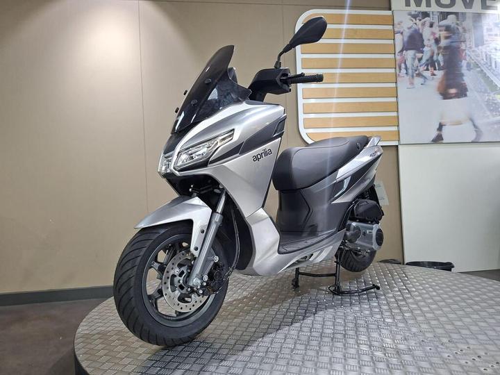 A silver Aprilia scooter displayed on a circular platform, set against a neutral background with a partially visible image of people in motion on the wall behind.