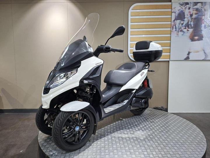 A modern three-wheeled scooter in white and black, featuring a large windshield and a rear storage box, displayed on a circular platform with a textured surface against a neutral background.