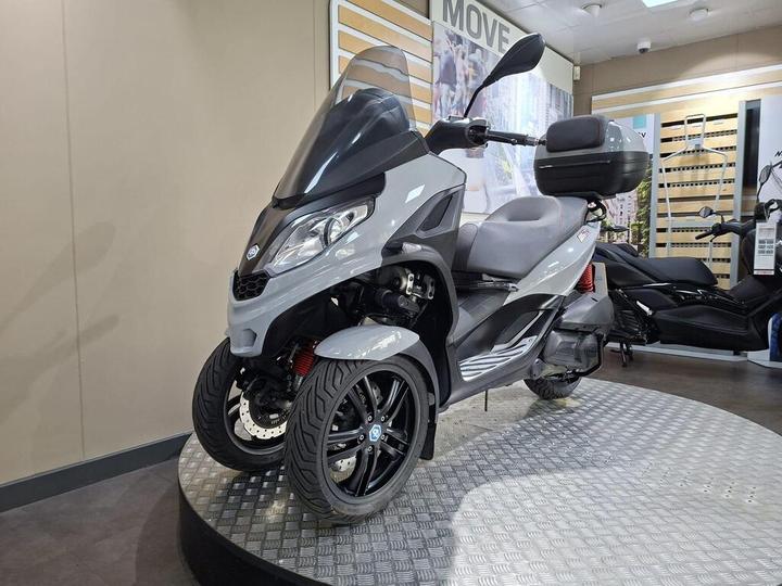 Three-wheeled gray scooter displayed on a circular platform in a showroom, featuring a large windshield, rear storage box, and modern styling.