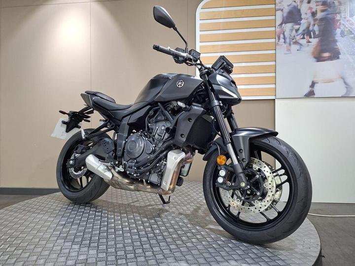 A black motorcycle is displayed on a textured platform against a beige wall with a promotional image in the background.