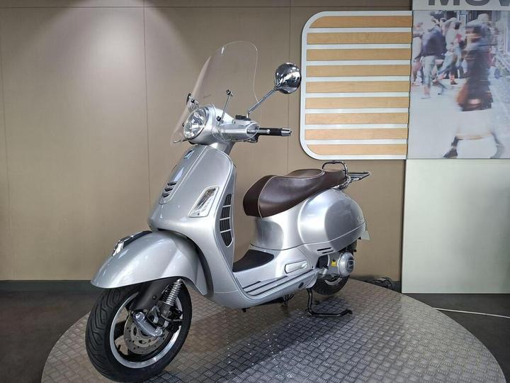 A silver scooter with a brown seat and a windshield, displayed on a circular platform against a neutral background with a graphic panel.