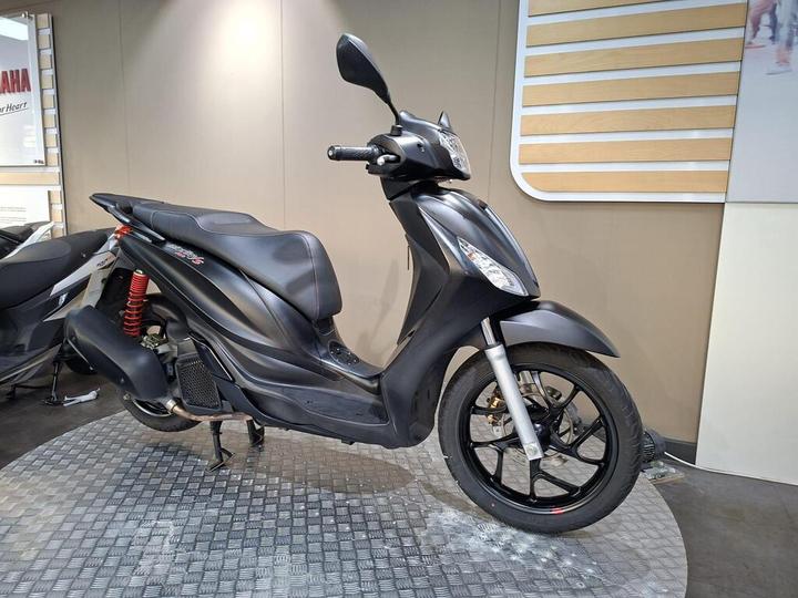 A sleek black scooter displayed on a textured circular platform in a modern showroom, featuring a sporty design and red shock absorbers.