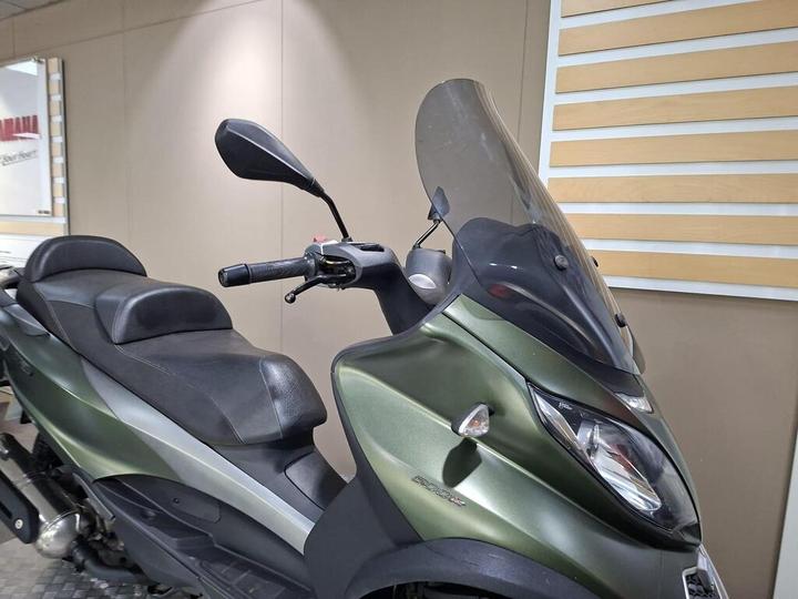 Side view of a green scooter with a large windshield, featuring a comfortable seat and handlebar controls, set against a neutral background.