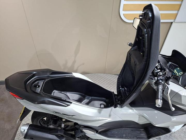 A close-up view of a motorcycle seat in an open position, revealing the storage compartment beneath. The bike features a sleek, modern design with a gray body and black accents.