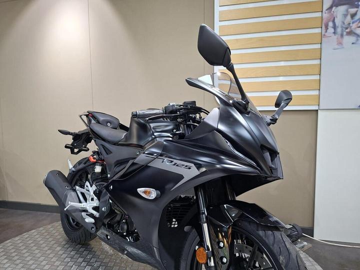 A sleek, matte black motorcycle displayed at an angle, showcasing its aerodynamic design, front fairing, and sporty features, set against a light-colored wall.