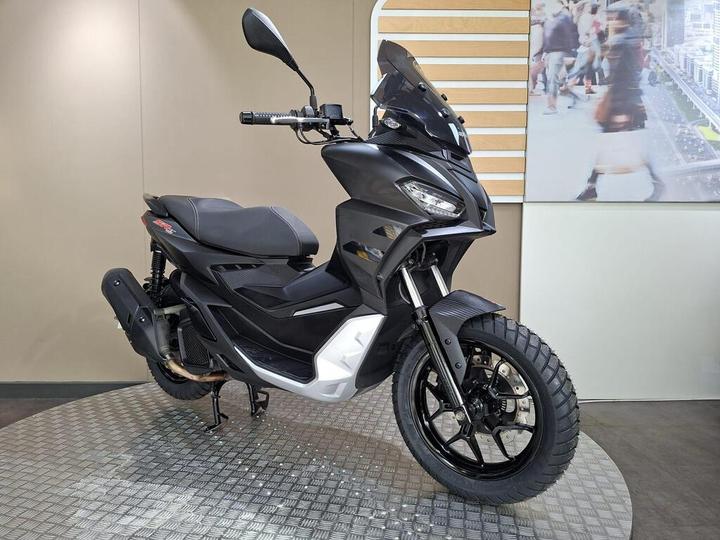 A sleek, black scooter displayed on a textured floor with a modern backdrop. The scooter features a sporty design with a prominent front fairing, off-road tires, and a high windshield.