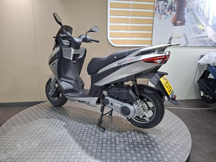 A sleek gray scooter displayed on a circular metal platform, with a backdrop of an indoor setting featuring modern design elements.