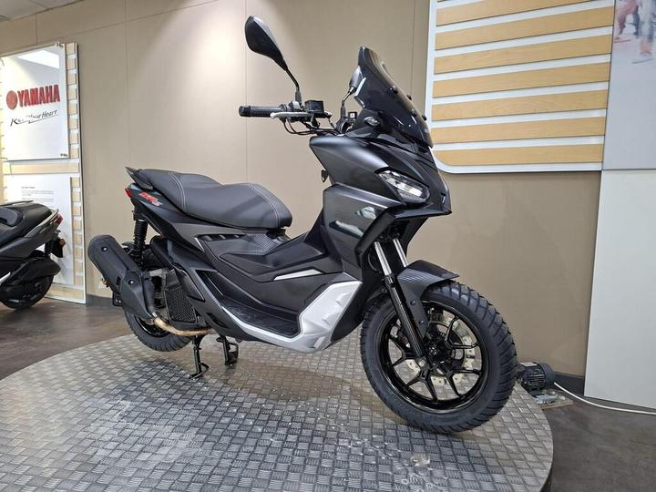 A sleek, black Yamaha scooter displayed in a showroom, featuring a modern design, off-road tires, and a stylish windscreen, with a light-colored patterned floor and minimalistic wall decor in the background.