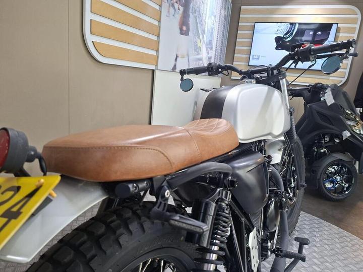A close-up view of a custom motorcycle featuring a brown leather seat, black frame, and a white fuel tank, with a modern digital display in the background.