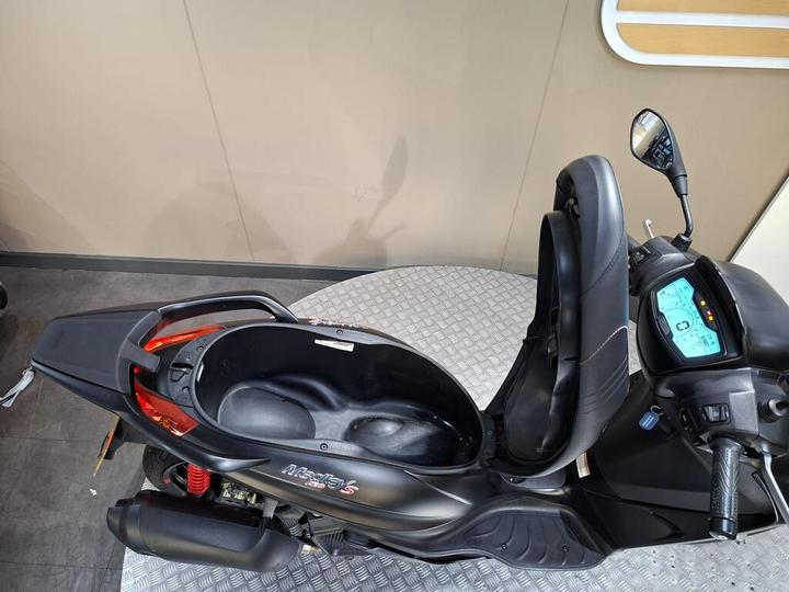 A black scooter sits on a gray textured platform, showcasing its seat and dashboard. The dashboard features a blue digital display with various indicators, while the scooter’s storage compartment is open, revealing its interior. The background is a neutral-colored wall.