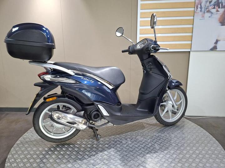 A dark blue scooter with a sleek design, featuring a black rear trunk, parked on a textured circular platform against a neutral background.