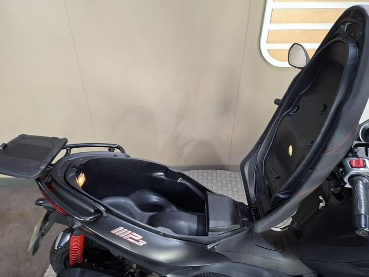 A close-up view of an open scooter's storage compartment, revealing a spacious interior with a light inside, and a removable top panel. The scooter features a black body and red suspension.