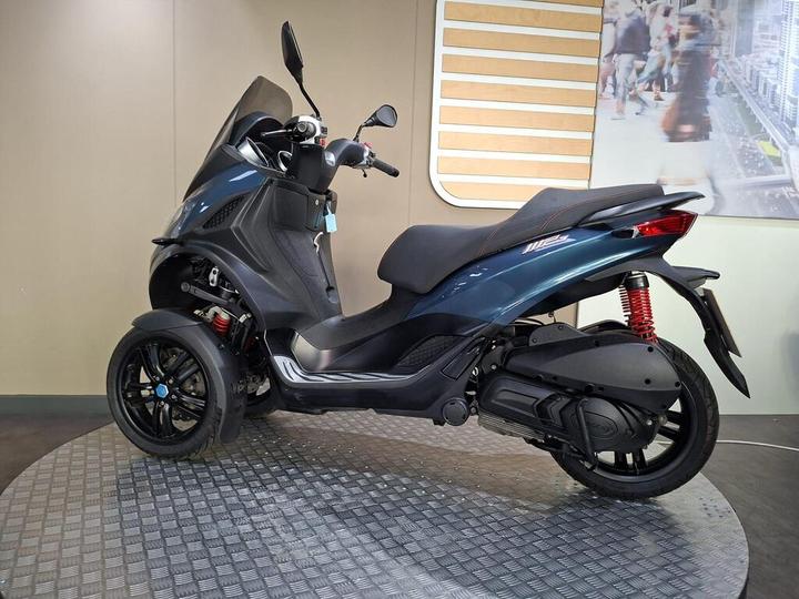 A side view of a blue three-wheeled scooter displayed on a circular platform, with a modern background featuring blurred people and a subtle design. The scooter has a sleek, sporty design with prominent red suspension and a black seat.
